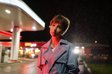 Sad lonely young woman with short haircut wearing grey trench coat standing against filling station...