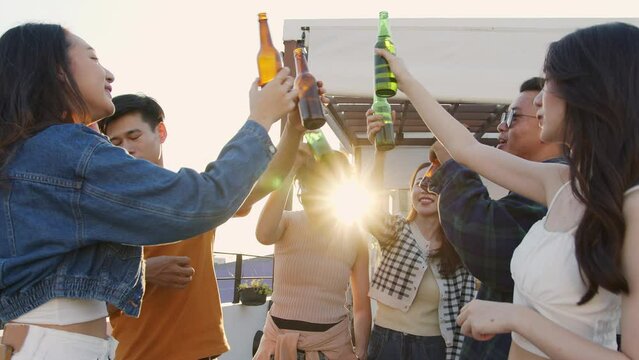 Group of young asian friends having fun drinking alcohol at sunset rooftop party. asian friends clinking with beer bottles on terrace in city background. 