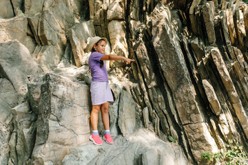 Fototapeta na wymiar Pretty child in panama hat stand on big rock and point finger at something interesting. Little girl in sports clothing and footwear walk and hike in mountains. Tourism, hiking, uphill climb, trip.