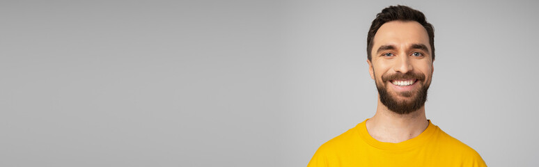 Fototapeta premium portrait of cheerful bearded man in bright yellow t-shirt looking at camera isolated on grey, banner.