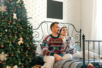 Obraz na płótnie Canvas Young woman and man sitting on bed in sweaters and hugging near Christmas tree