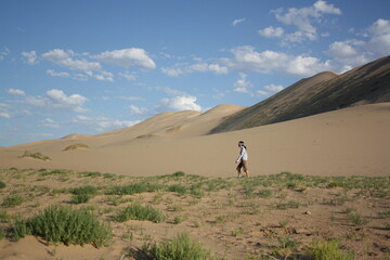 Fototapeta na wymiar The woman walks between the green vegetation and Khongor sand dunes in Gobi Desert, Umnugovi province, Mongolia. The sand dunes are so vast and windy. 