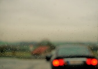 Glasscheibe mit Regentropfenmuster vor Landstraßenkreuzung mit zwei Autos, Ackerfeld, Wald und diesigem grauen Himmel bei Regen am Morgen im Herbst