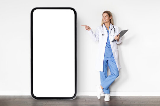 Smiling Doctor Woman In Uniform Pointing Finger At Big Blank Smartphone Screen