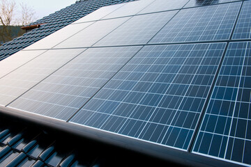 New solar panels are mounted on a roof with shiny black tiles and the clear sky is reflected in the...