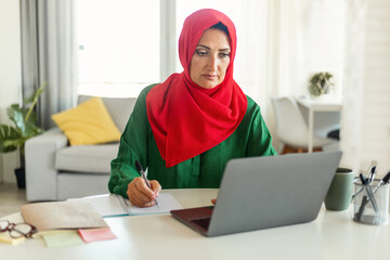 Focused muslim woman in hijab using laptop and calculating budget, sitting at table in living room...