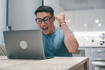 One young man working and winning something with laptop at home. Happy and euphoric teenager...