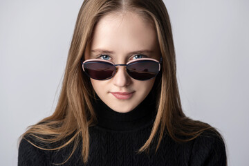 Make-up, beauty and fashion concept. Close-up portrait of brunette woman with sunglasses looking through glasses direct to camera and wearing black long neck sweater in grey studio background