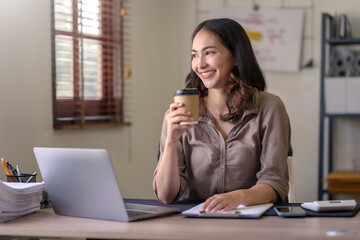 Beautiful Asian businesswoman sitting at her laptop happily working on her laptop and drinking coffee with a smile and looking at the camera looking bright in the office.