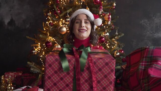 Woman wearing red sweater and Santa's cap presenting a gift near the tree