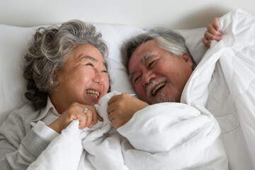 Top view of happy smiling asian elderly couple hugging and sleeping together on bed with white blanket, pillow in the bedroom at home. Retirement, health care, relax and spending time concept
