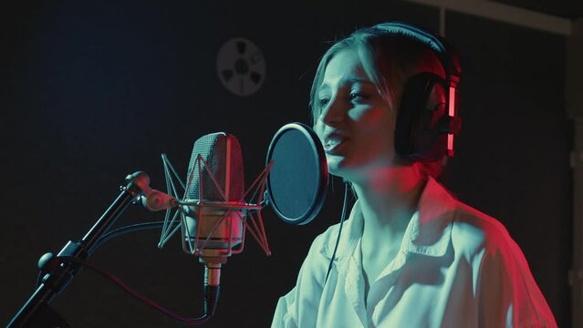 Beautiful blonde girl in headphones performs song in recording studio. Young woman sings into microphone, dances, enjoying rhythm in sound room. Singer is working on her new music album