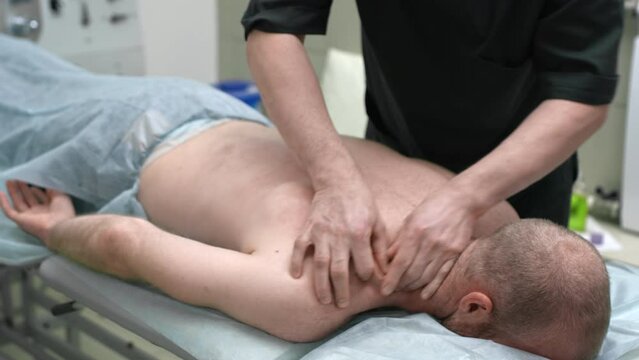 A doctor in a medical office massages a patient. Therapeutic massage after sports exercises. The therapist kneads the joints and muscles on the man's back with his hands. Body care
