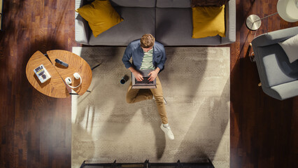 Top View Apartment: Young Man Uses Laptop Computer Sitting on a Carpet in Living Room. Creative...