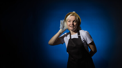 A woman in a white t-shirt, black apron, gloves and a hat put her hand to her ear and eavesdrops on...