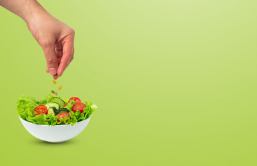 White bowl of salad with fresh cucumbers, tomatoes, iceberg salad leaves and male hand sprinkling pumpkin seeds over salad isolated on green background, template for advertising, space for text