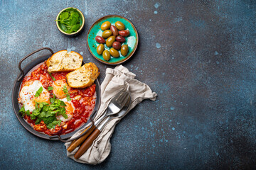 Middle Eastern and Maghrebi healthy dish Shakshouka made of eggs and tomato sauce served in pan with toasts, fresh cilantro and olives on rustic concrete background table from above, space for text