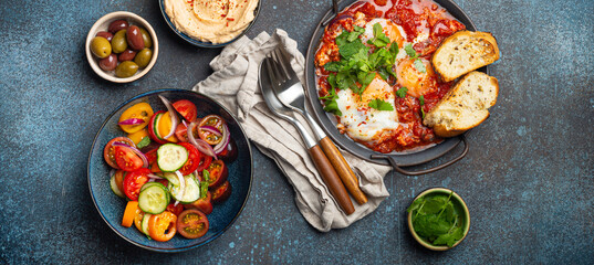 Middle Eastern traditional breakfast or brunch with eggs Shakshouka in pan with toasts, fresh vegetables salad, hummus and olives on rustic concrete background table from above - obrazy, fototapety, plakaty
