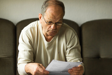 Worried senior man in white sweater and glasses reading utility bill in period of energy crisis....