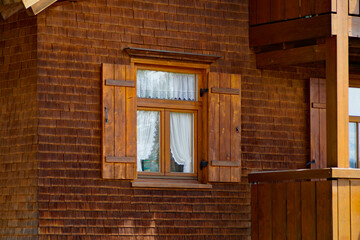 Obraz na płótnie Canvas a beautiful rustic window with wooden shutters and white lacy drapes (Schuppenflechtehouse) in the alpine village in Ofterschwang in the Bavarian Alps on a sunny spring day (Bavaria, Germany)