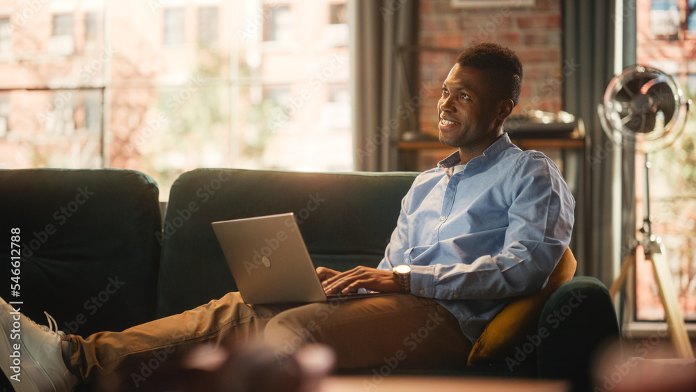 Poster handsome african american man working on laptop computer while sitting on a sofa in cozy living room