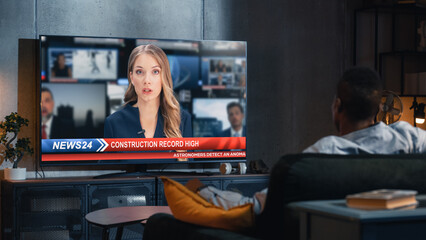 Over the Shoulder View of Black Man Watching TV News, Sitting on Couch in Stylish Loft Living Room. TV Presenter Telling Breaking News About Construction Industry. Home with Cozy Interior.
