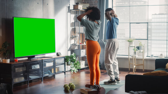 Athletic Black Couple Training Together, Jumping Infront of Green Screen Chromakey Placeholder on TV Display in Bright Sunny Room at Home. Beautiful Boyfriend and Girlfriend Workout Together.
