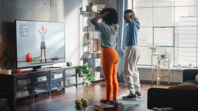 Athletic Black Couple Training Together, Jumping, Following Online Video Exercise from Professional Trainer on TV in Bright Sunny Room at Home. Beautiful Boyfriend and Girlfriend Workout Together