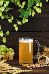 Beautiful unfiltered beer with foam in a glass glass with hop cones and wheat ears on a wooden background, vertical arrangement