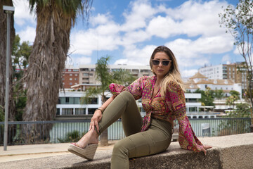 Young, attractive and blonde woman, wearing cashmere shirt and green trousers, sitting on a stone wall, on vacation. Concept beauty, fashion, trend, holidays, travel.