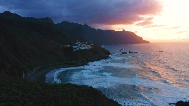 Aerial flight over deep dark night ocean water. Sea waves crash on rocky beach. Last rays of sunset on cloudy colorful sky. Little village Almaciga on top of mountain. Tenerife north. Canary island.