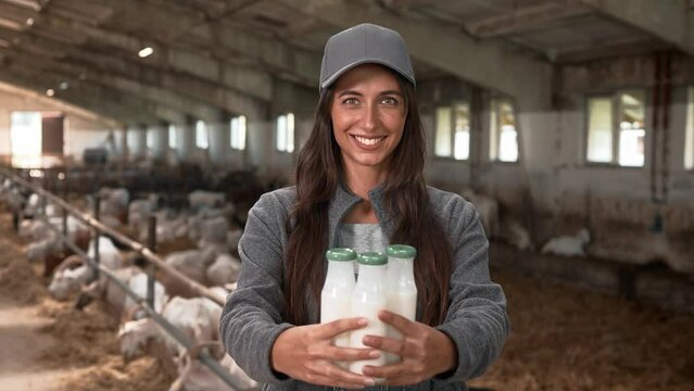 Close up of Caucasian happy young woman holding in hands bottles with milk standing in barn indoor. Beautiful female farmer giving fresh ecological goat milk and smiling at camera. Animal farming