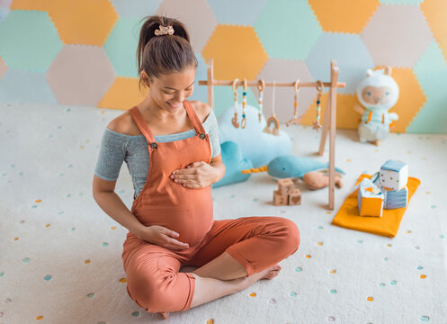 Pregnant woman smiling at belly in nursery playrooom with Baby activity gym play, toys and playmat. Pregnancy concept and home nusery planning and decoration concept photo with happy expecting woman