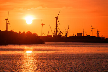 Industrial buildings along with wind turbines and dock cranes on a river harbour silhouetted...