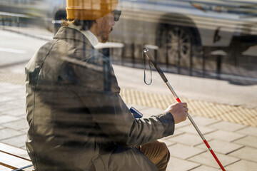 Blind man with a walking stick sitting on a bench at the tram stop behind glass