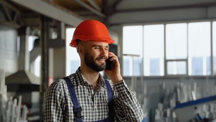 A young worker in a helmet at a large metalworking plant speaks on the phone. Professional worker in the finished product warehouse makes an order