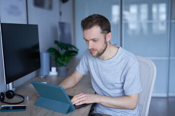 Attractive bearded male sitting in home office using tablet working or educating online. Close-up of portrait of young handsome man in grey shirt. High quality photo