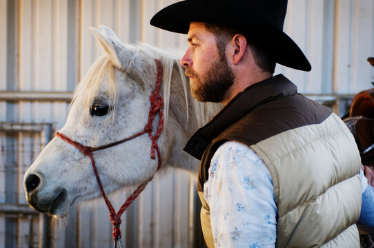 A male ranch hand in a cowboy hat strokes the neck of Nell, a four year old arabian mare, and gets ready to go out on a horseback ride in Flagstaff, AZ.