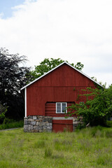 Fototapeta na wymiar Big red farmer's barn in Norway with gray foundations of big stones, green grass and some green bushes grow around the barn. Above the barn is a sky of bright poppies.