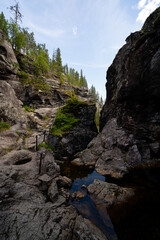 blue dark narrow valley in Norway, Helvete, flanked by a forest with green trees and a blue sky with puffy clouds above them and a metal railing along the edge of the cliff