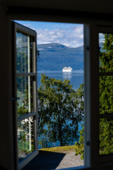 an open window of a small house, through which you can see green trees, behind which is the blue water of the fjord, through which a large white cruise ship floats