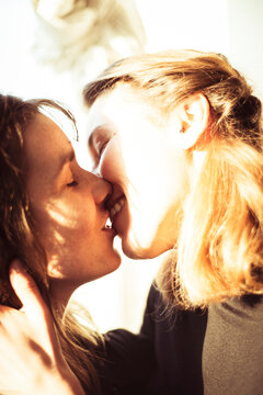 Feminine lesbian couple kiss and smile in warm sunlight in window