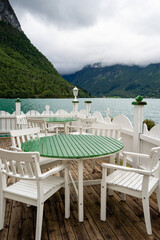 A brown terrace with a white wooden railing where there are white chairs with round green tables and in the background behind the terrace green fjord water with green mountains behind