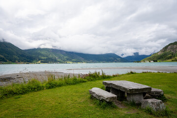 a stone table and benches stand in the grass by the blue fjord, which is a mountain caught in gray...