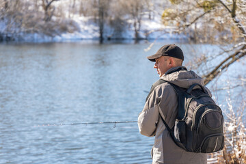 A fisherman with a fishing rod and a backpack catches fish on the bank of a snow-covered river in early spring