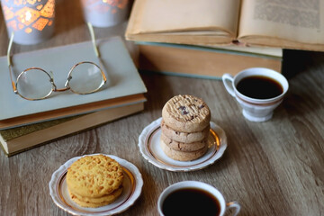 Plates with various cookies, cups of tea or coffee, vintage books, reading glasses and lit candles on the table. Selective focus.