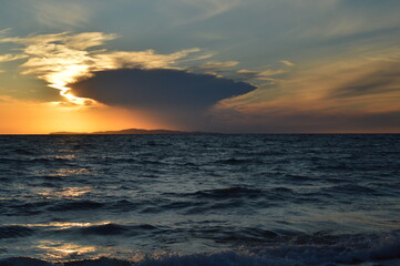 A dramatic sunset behind a small island in the distance with a large storm cloud above it	