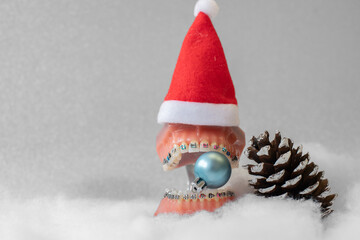 
Lustiger zahnärztlicher Weihnachtshintergrund mit Platzhalter. Zahnmodell mit festsitzender Zahnspange trägt eine Weihnachtsmütze und isst eine Weihnachtskuge - obrazy, fototapety, plakaty