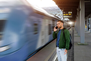 Caucasian man traveler in green windbreaker and with backpack stands on platform talking on the phone, electric commuter train motion speed blur passes by in morning at station. © aapsky