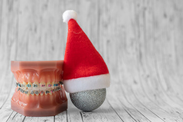 
Lustiger zahnärztlicher Weihnachtshintergrund mit Platzhalter. Zahnmodell mit festsitzender Zahnspange trägt eine Weihnachtsmütze und isst eine Weihnachtskugel - obrazy, fototapety, plakaty